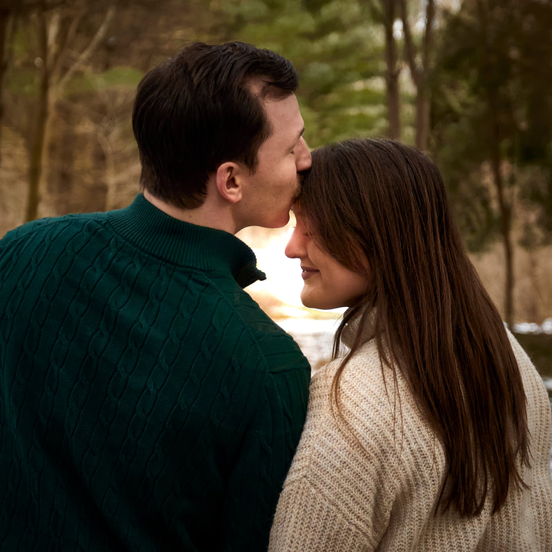 A man kissing his fiancé on the forehead in the woods
